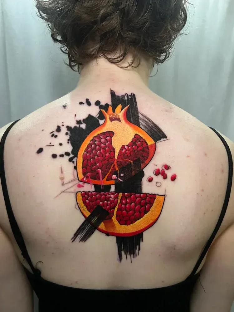 A back tattoo of a pomegranate sliced in half, rendered in vibrant red and orange with bold black ink brushstroke accents.