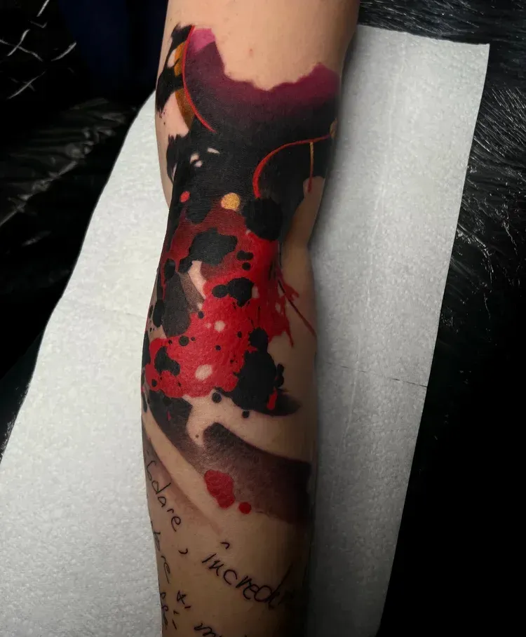 A black and red abstract watercolor-style tattoo on an arm, featuring ink splatters and a section of handwritten text.