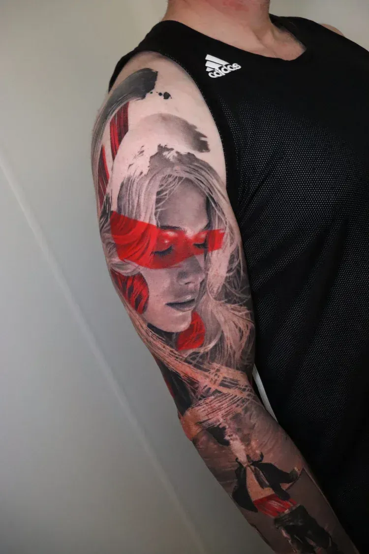 A tattoo on a person's upper arm featuring a portrait with red painted strokes across the eyes and a figure below.