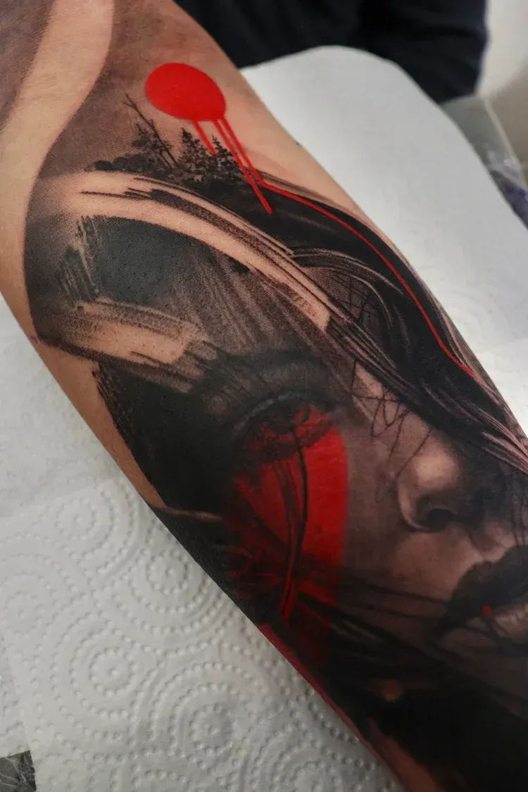 A tattoo on a forearm featuring a stylized portrait with abstract black brushstrokes and bold, dripping red accents.