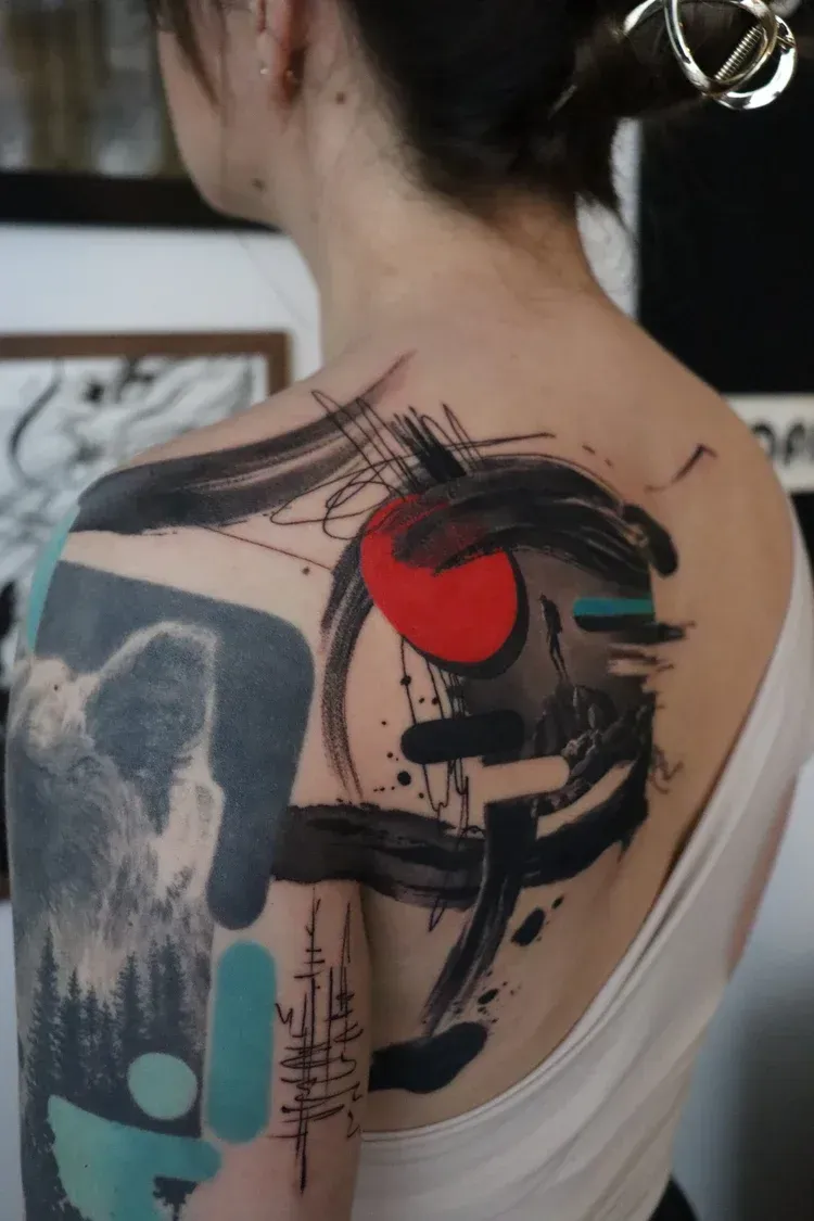 Abstract tattoo on a shoulder featuring bold black brushstrokes, a bright red circle, and teal geometric accents.