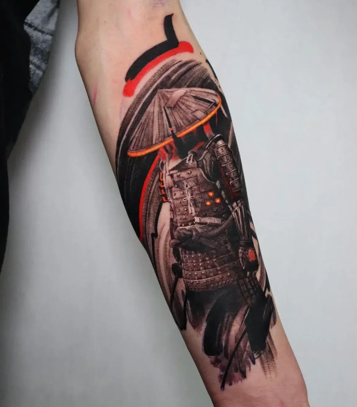 A forearm tattoo of a samurai wearing a wide hat and armor, rendered in black and grey with bold red and orange accents.
