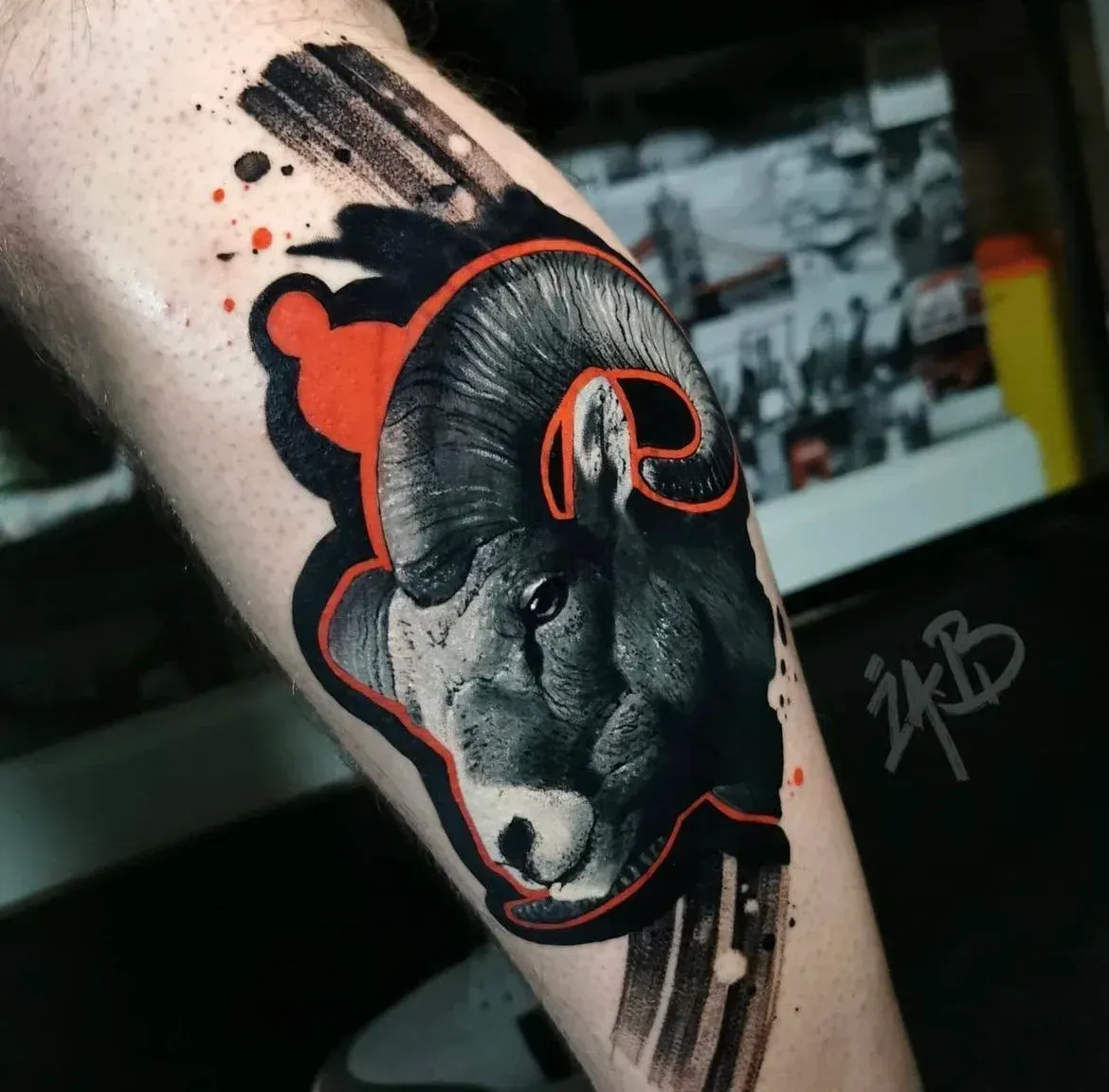A detailed tattoo of a bighorn sheep on an arm, featuring black ink shading and bold red outlines with abstract strokes.