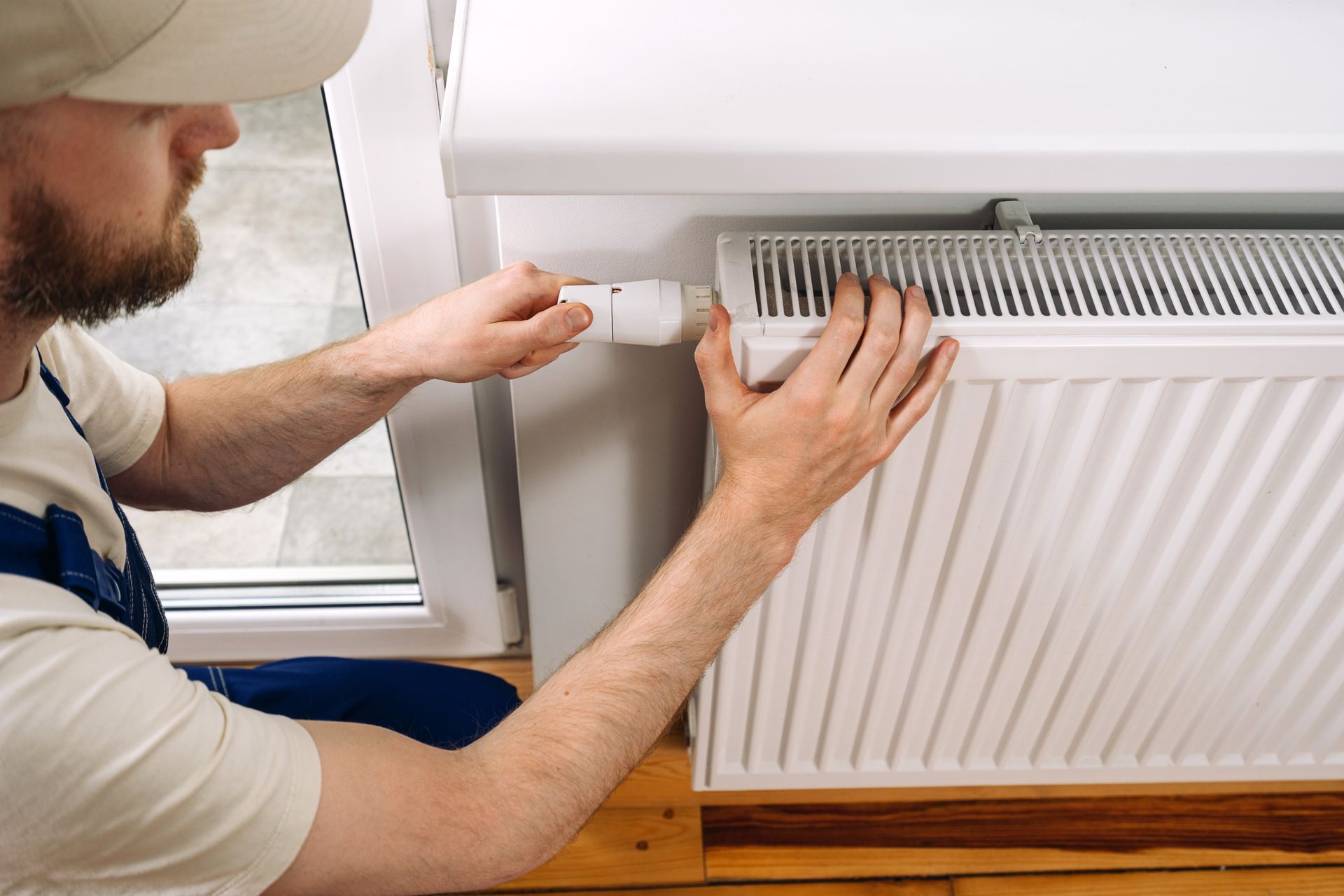 Man in overalls and cap adjusting a white radiator valve near a window.
