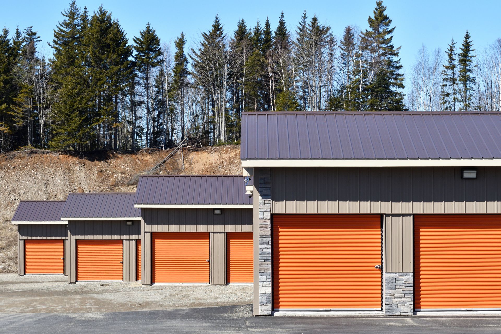 Orange storage unit doors with gray roofs and siding against a backdrop of trees and a blue sky.