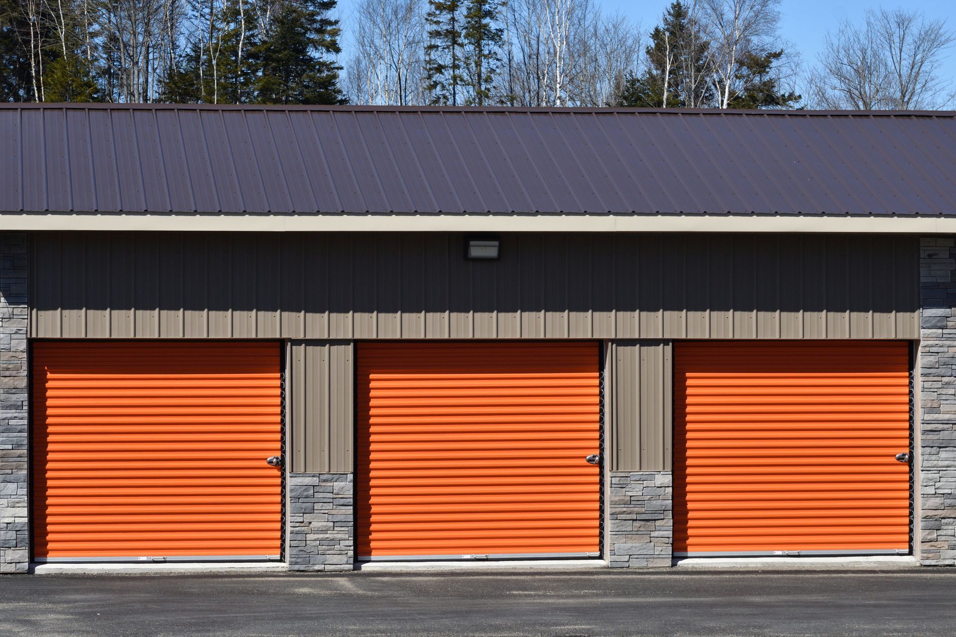 Three orange storage unit doors beneath a brown metal roof and gray siding.