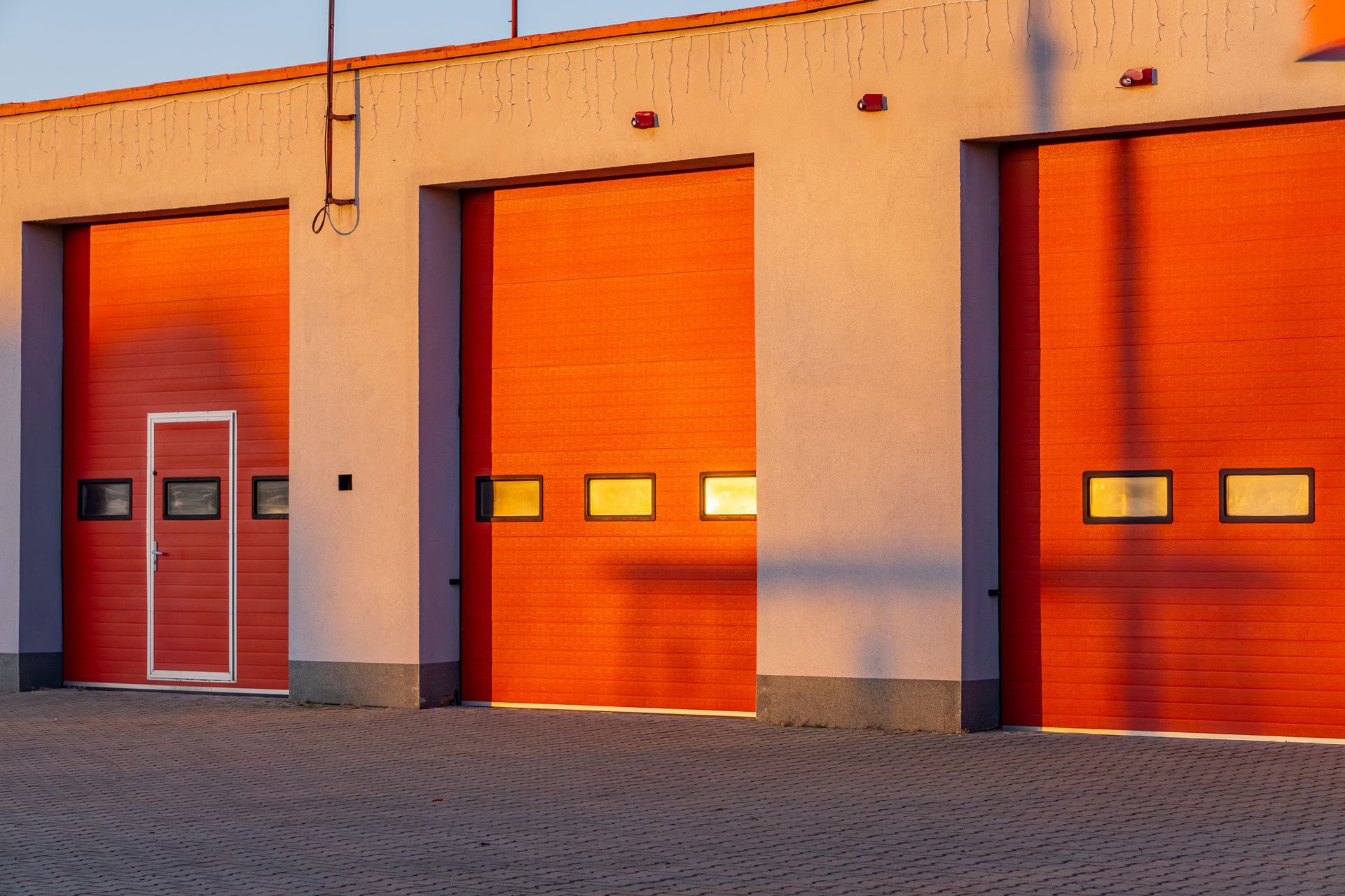 Three orange garage doors in a building exterior, lit by the setting sun. A white door is on the left, along with small windows on two doors.