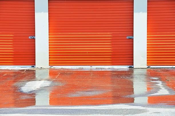 Three orange storage unit doors with white columns reflected in a wet, reddish pavement.