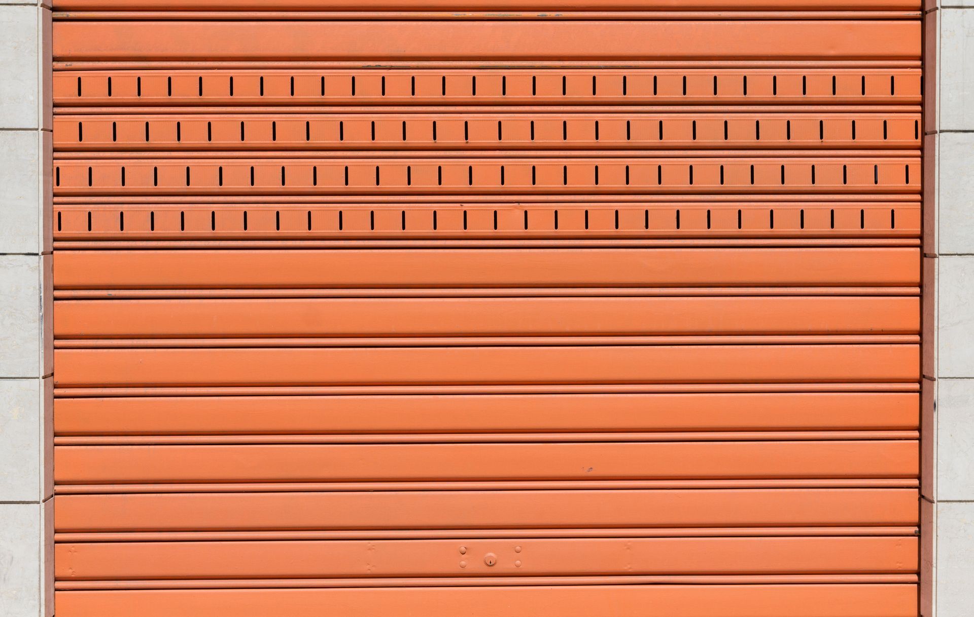 Orange metal rolling shutter with ventilation slits, set between light-colored tiled walls.