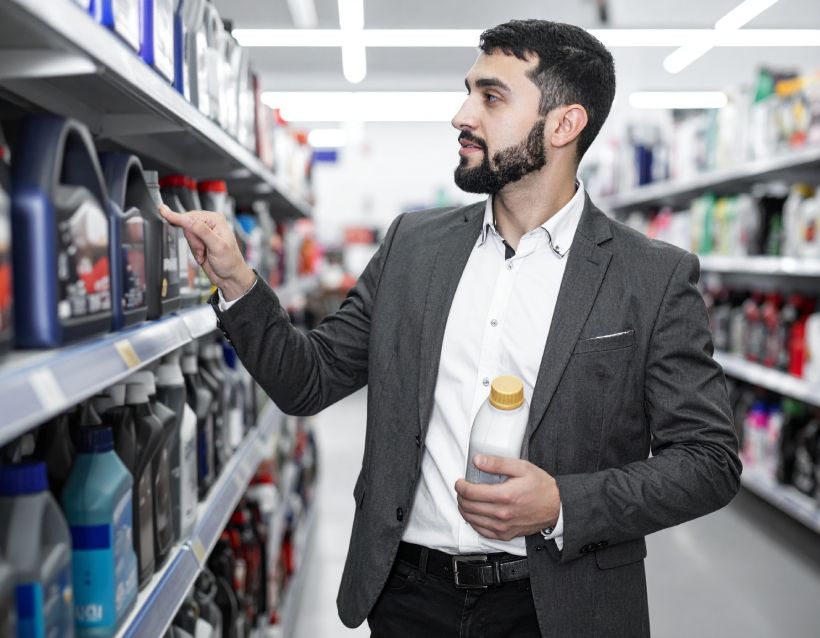 Man in a suit looking at motor oil bottles in a store. Holds a white bottle.