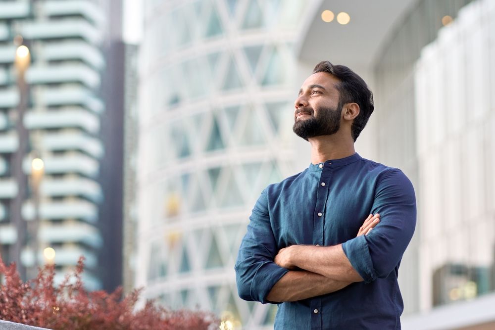 A man with a beard is standing in front of a building with his arms crossed.