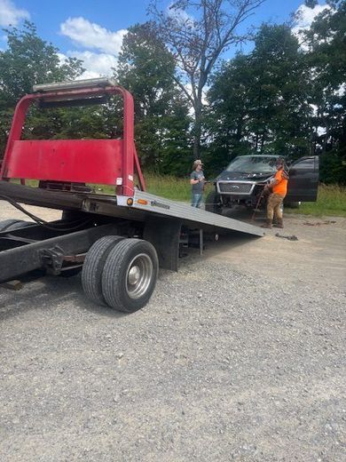 Tow truck loading a dark pickup truck. Two people stand near the truck on a gravel lot.
