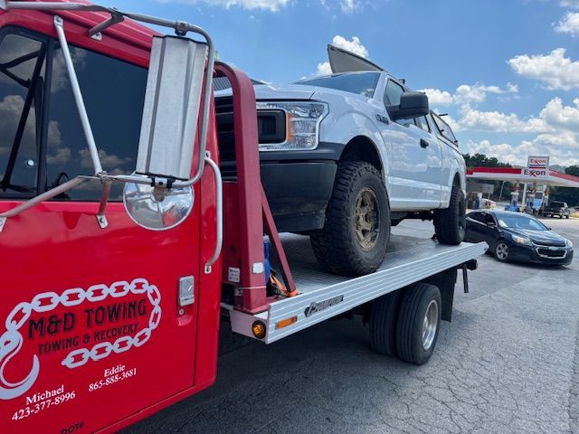 Red tow truck carrying a white Ford truck on a flatbed, near a gas station on a sunny day.