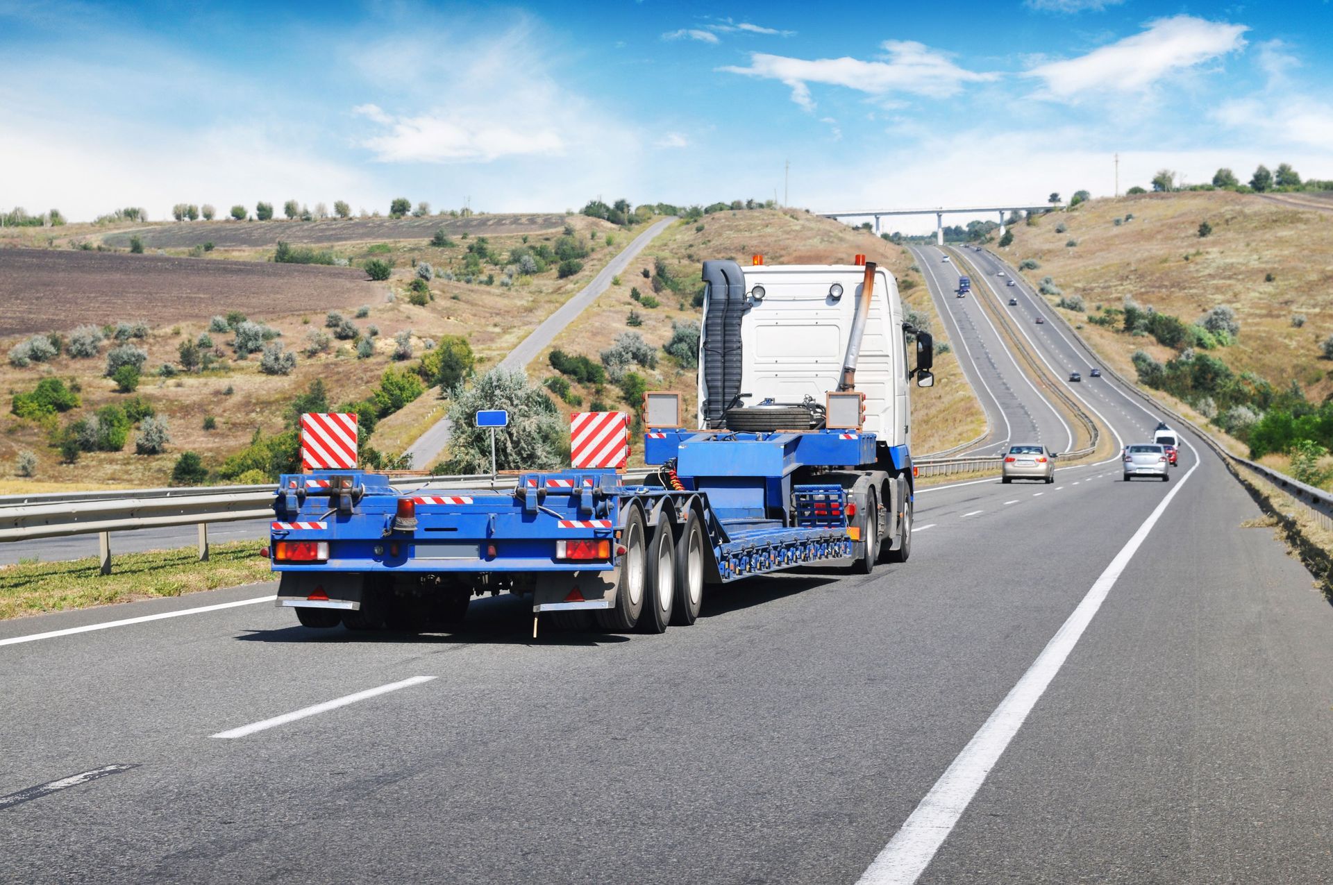 Blue and white semi-truck with empty trailer on highway, rolling hills in background, sunny day.