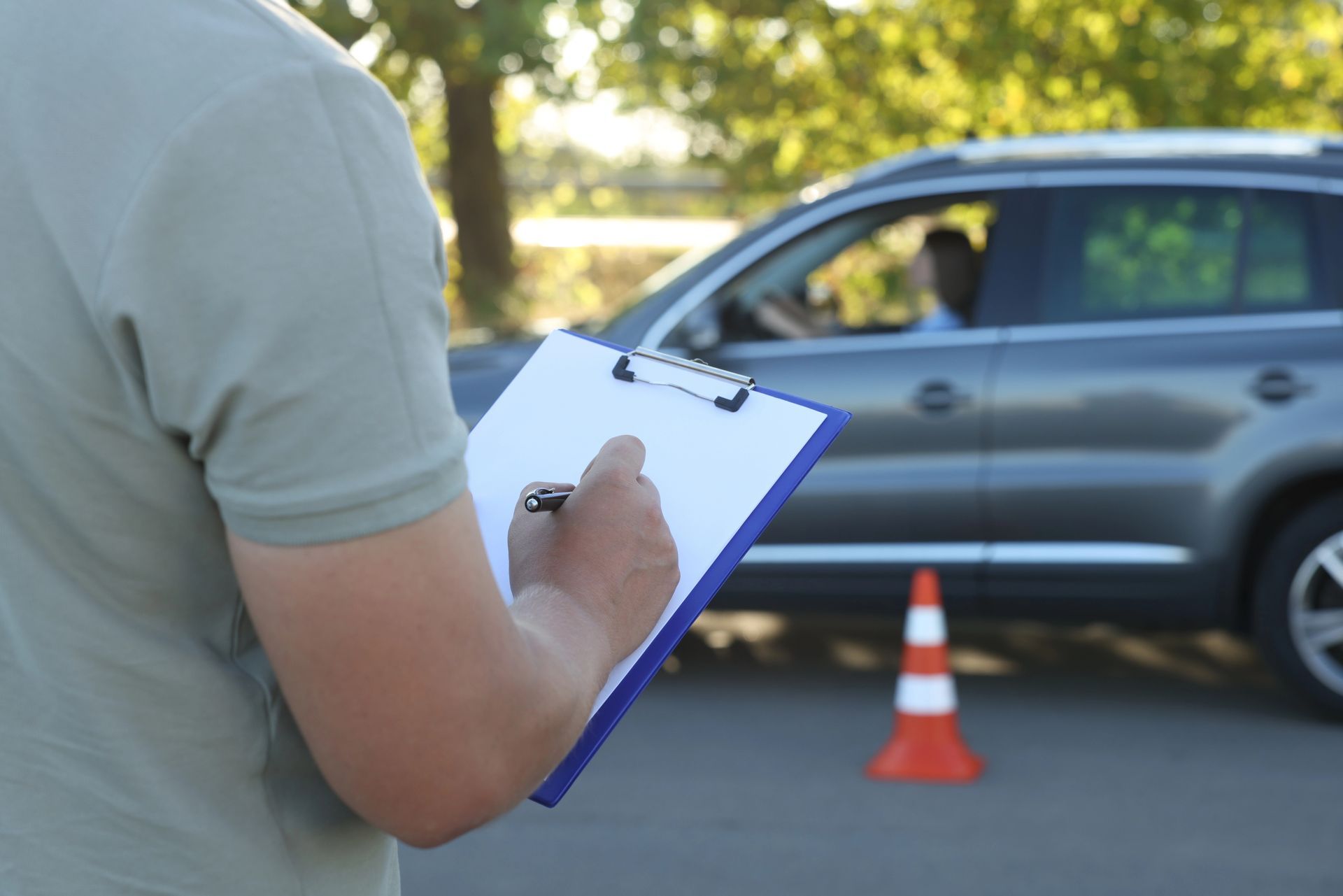 Instructor de manejo evaluando a un conductor, sosteniendo un portapapeles, al lado de un auto gris y un cono de tráfico.