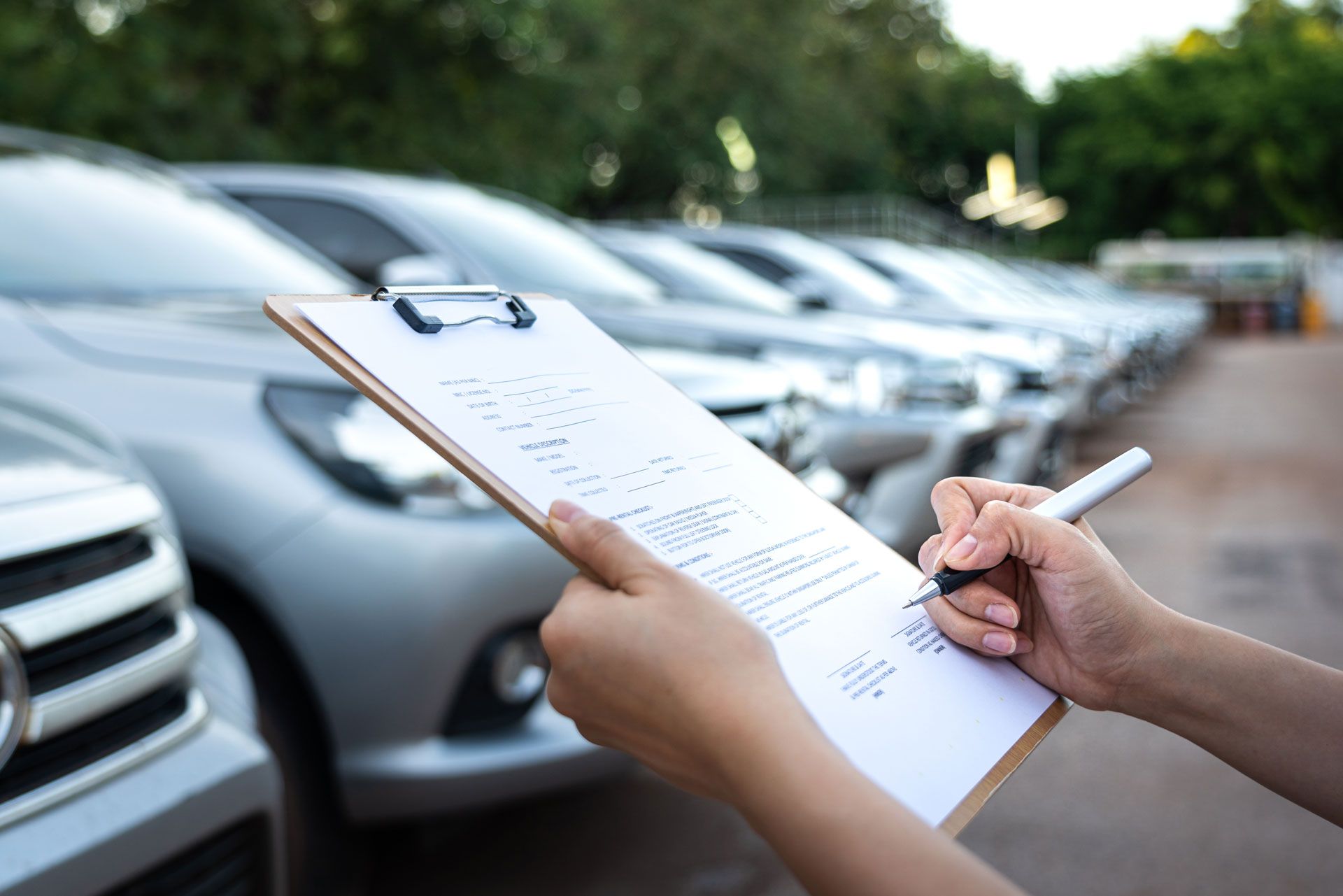 Person writing on clipboard in front of a row of silver cars.