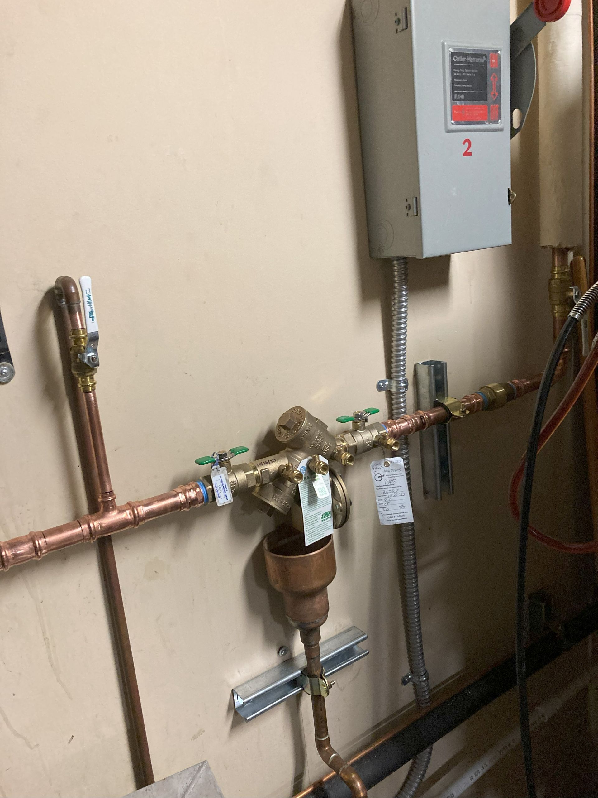 Water meter and plumbing pipes mounted on a wall with an electrical panel above.