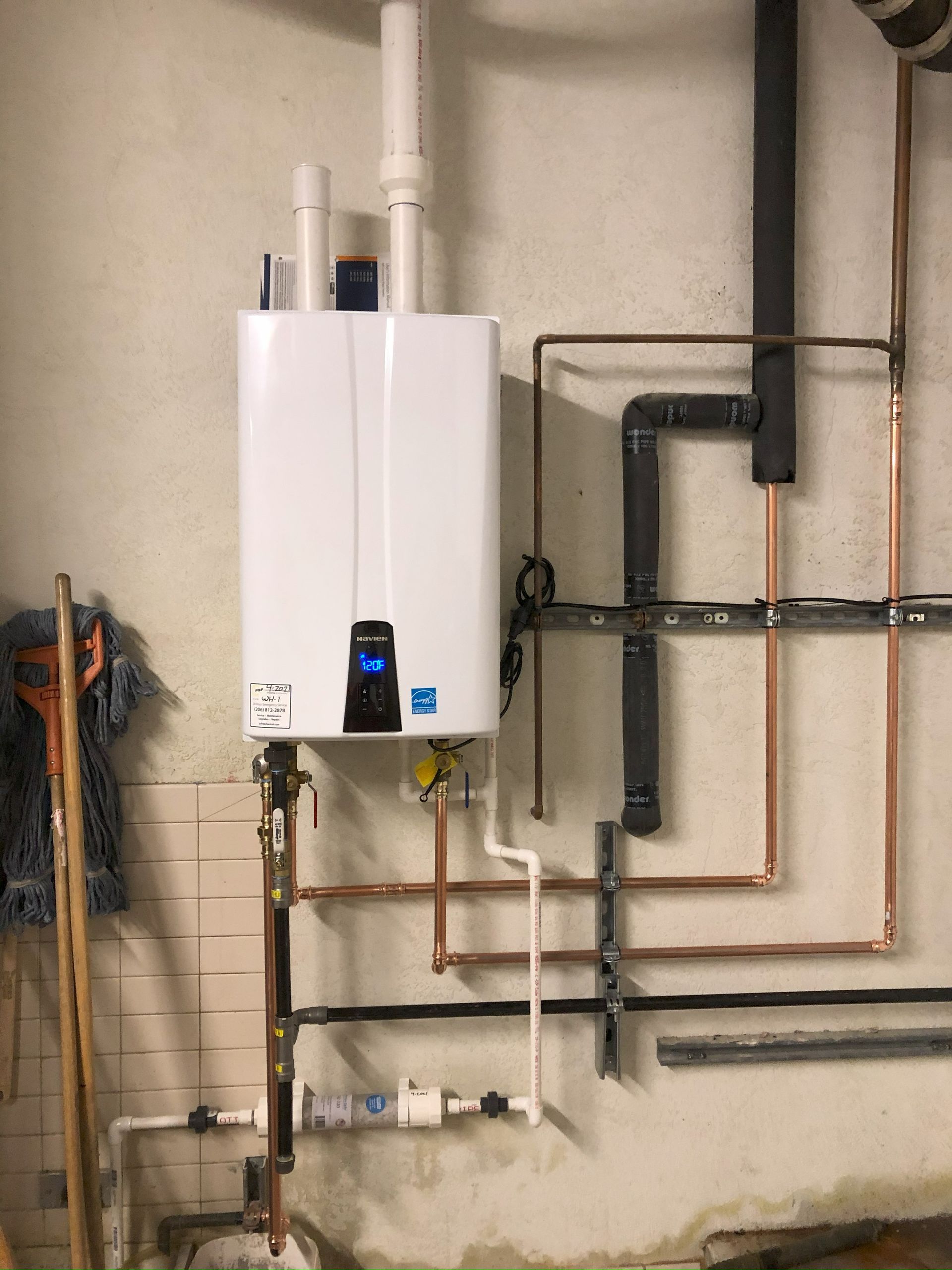 White wall-mounted boiler with exposed pipes and valves on a basement wall