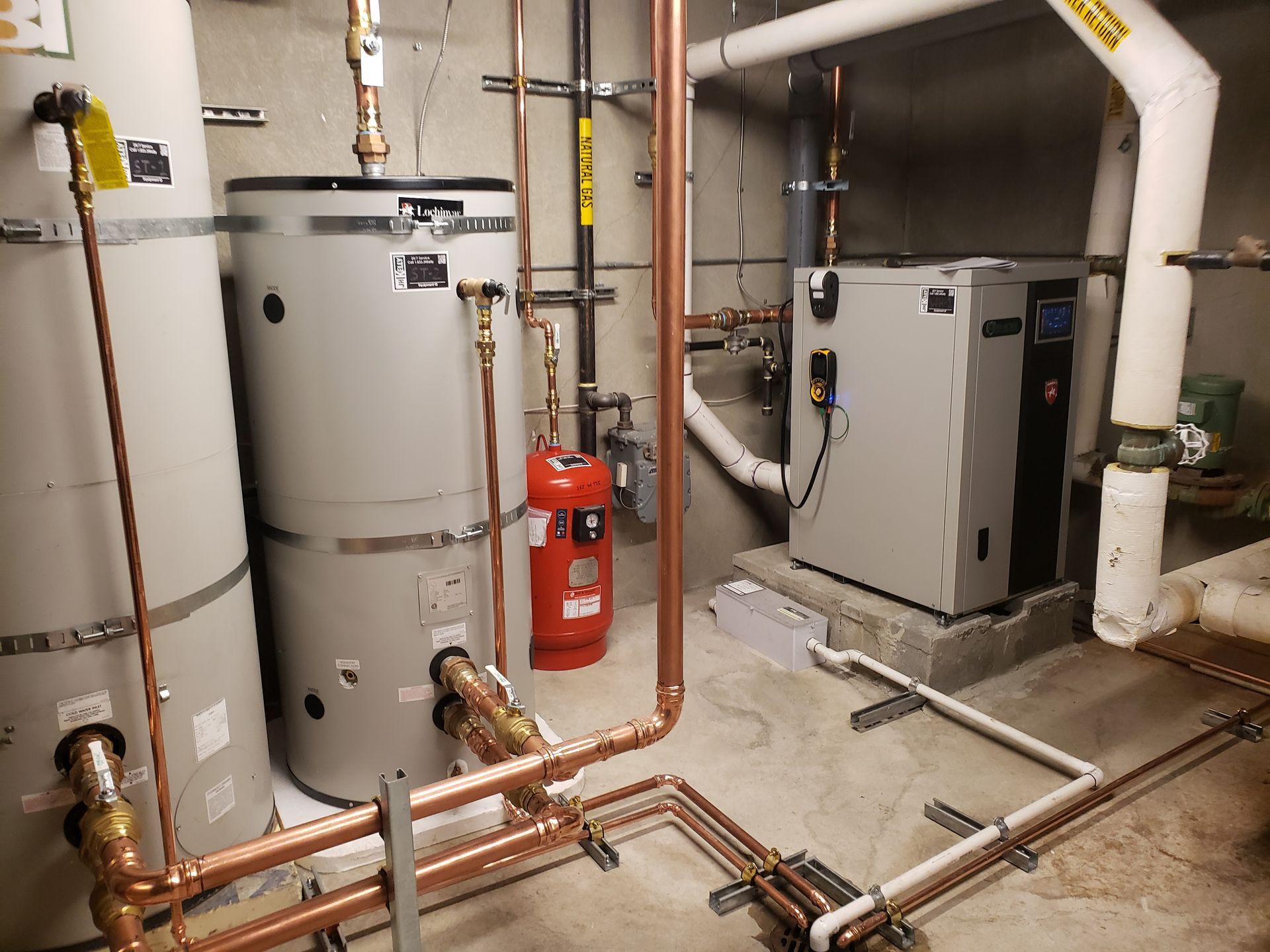 Mechanical room with boilers, pipes, valves, and a red fire extinguisher on the wall.