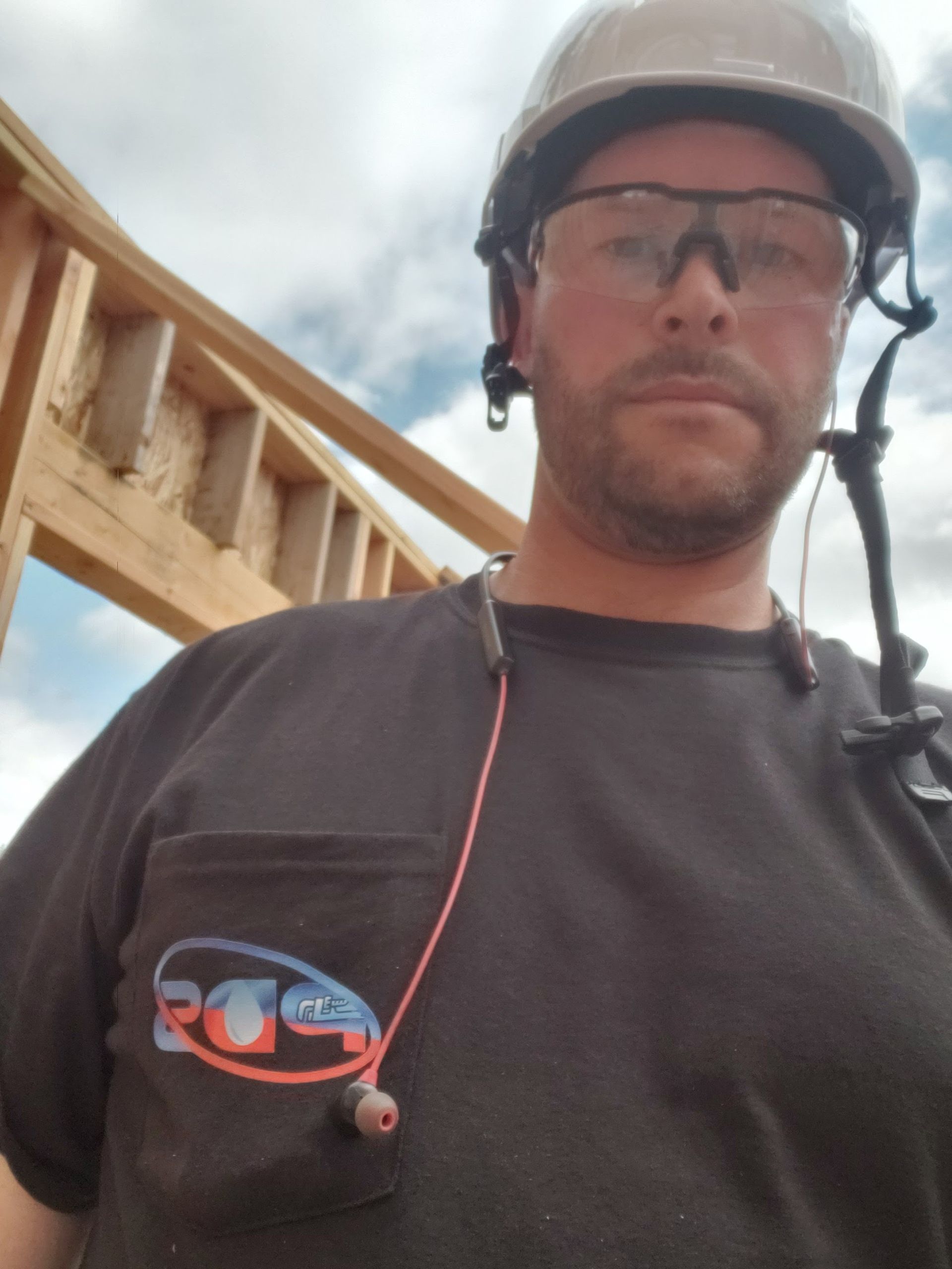 Construction worker wearing a hard hat and safety glasses at a building site, with wooden framing behind him