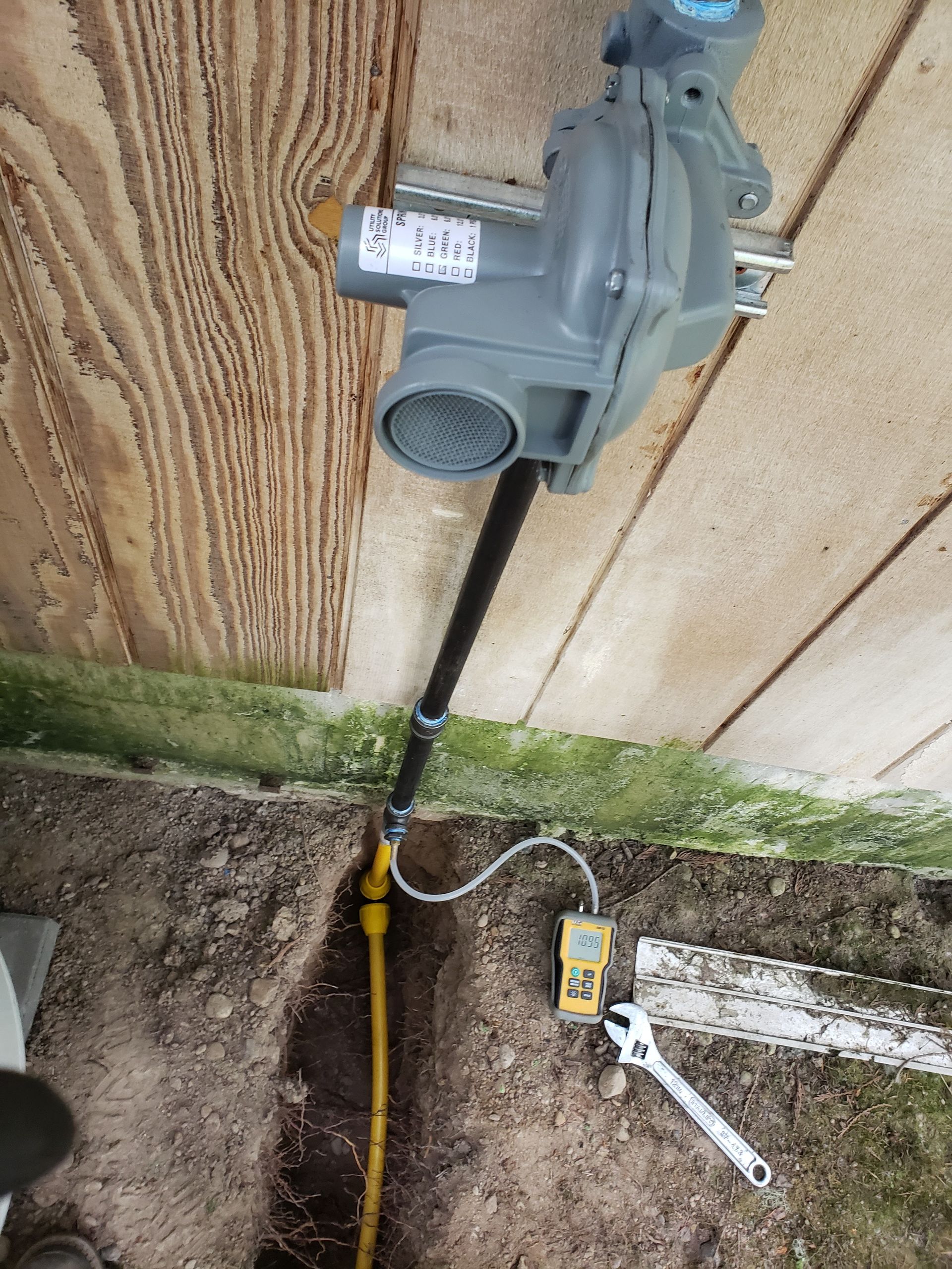 Water meter and shutoff valve beside a wooden wall, with a yellow hose and tools on the ground.