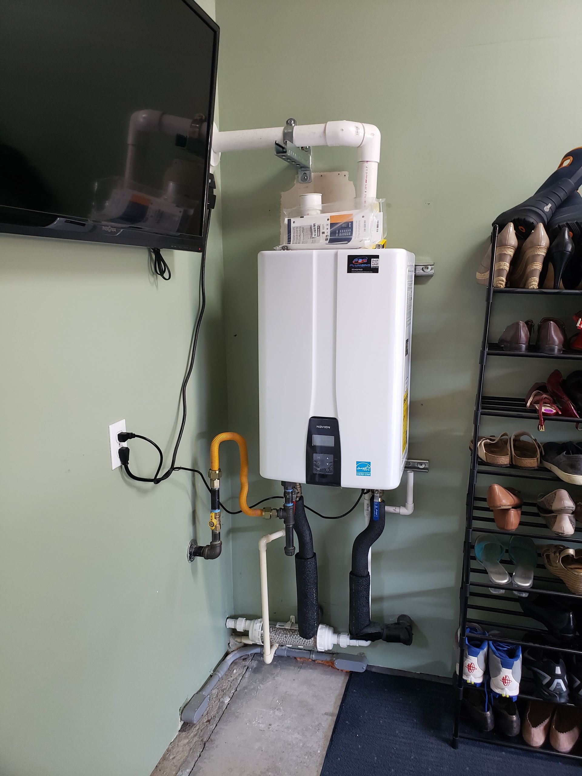 Wall-mounted water heater with exposed pipes beside a TV and shoe rack in a small room.