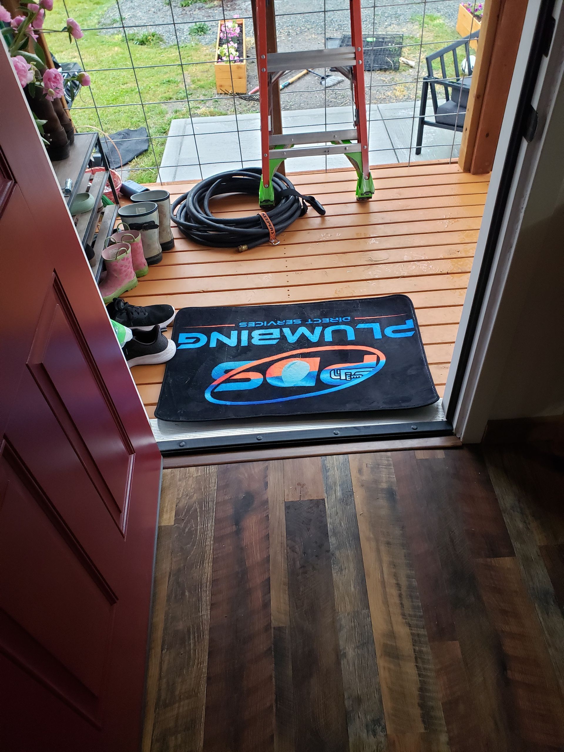 Open door to a small porch with a dark “Welcome” mat, ladder, and coiled hose on wooden decking.