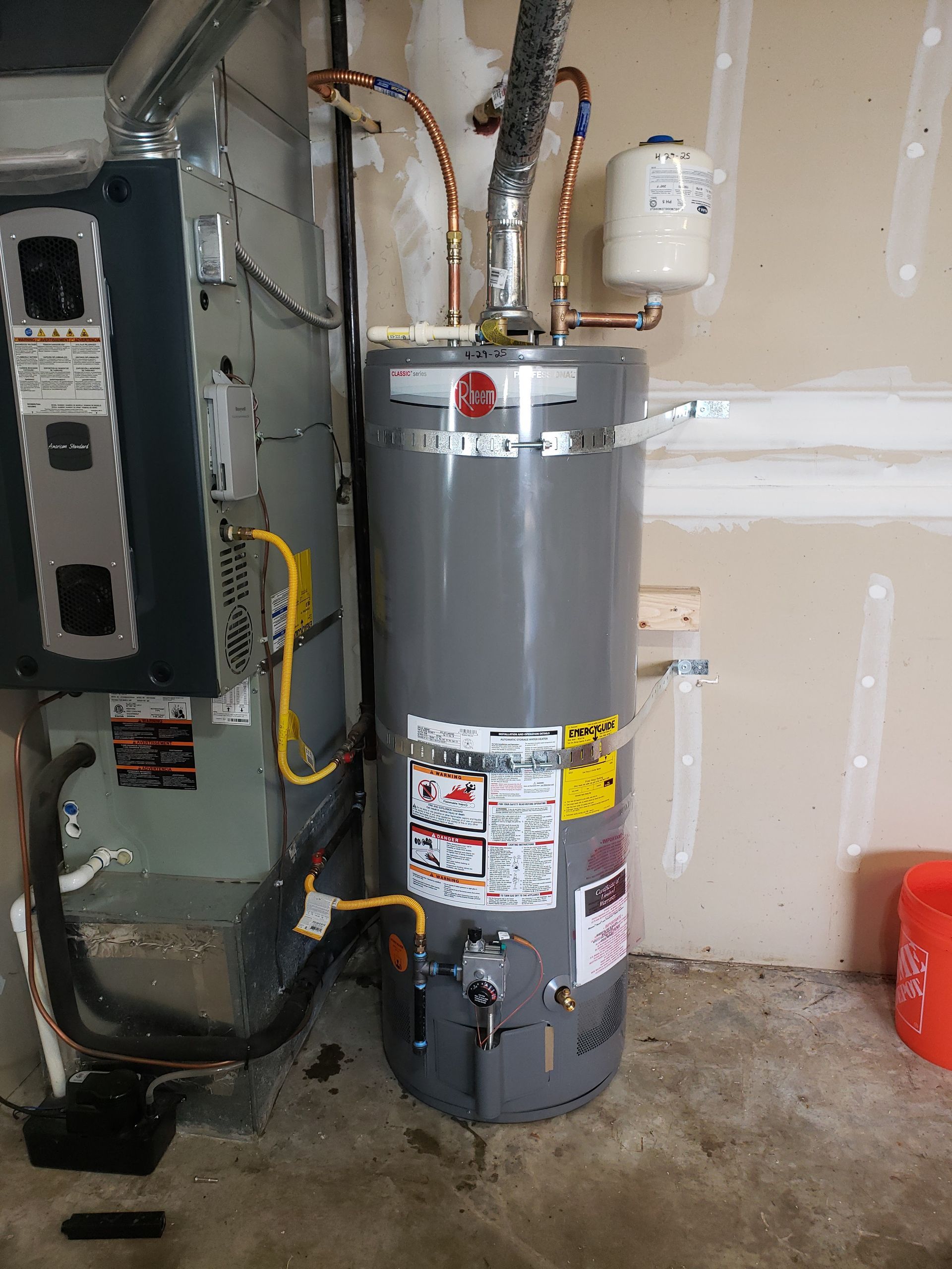 Gray water heater in a garage next to an electrical panel and exposed utility pipes.