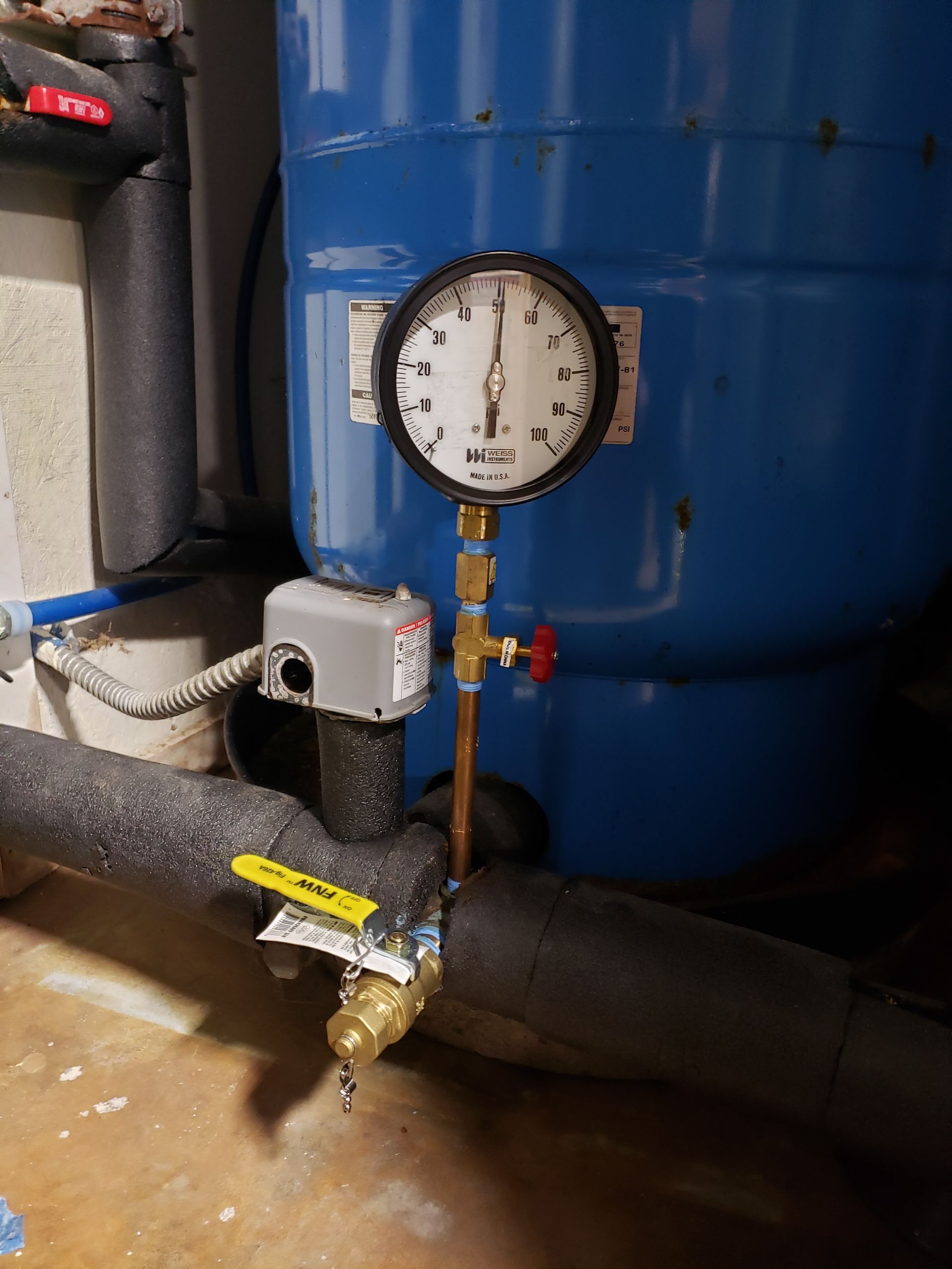 Blue industrial tank with pressure gauge, valves, and piping in a utility room.
