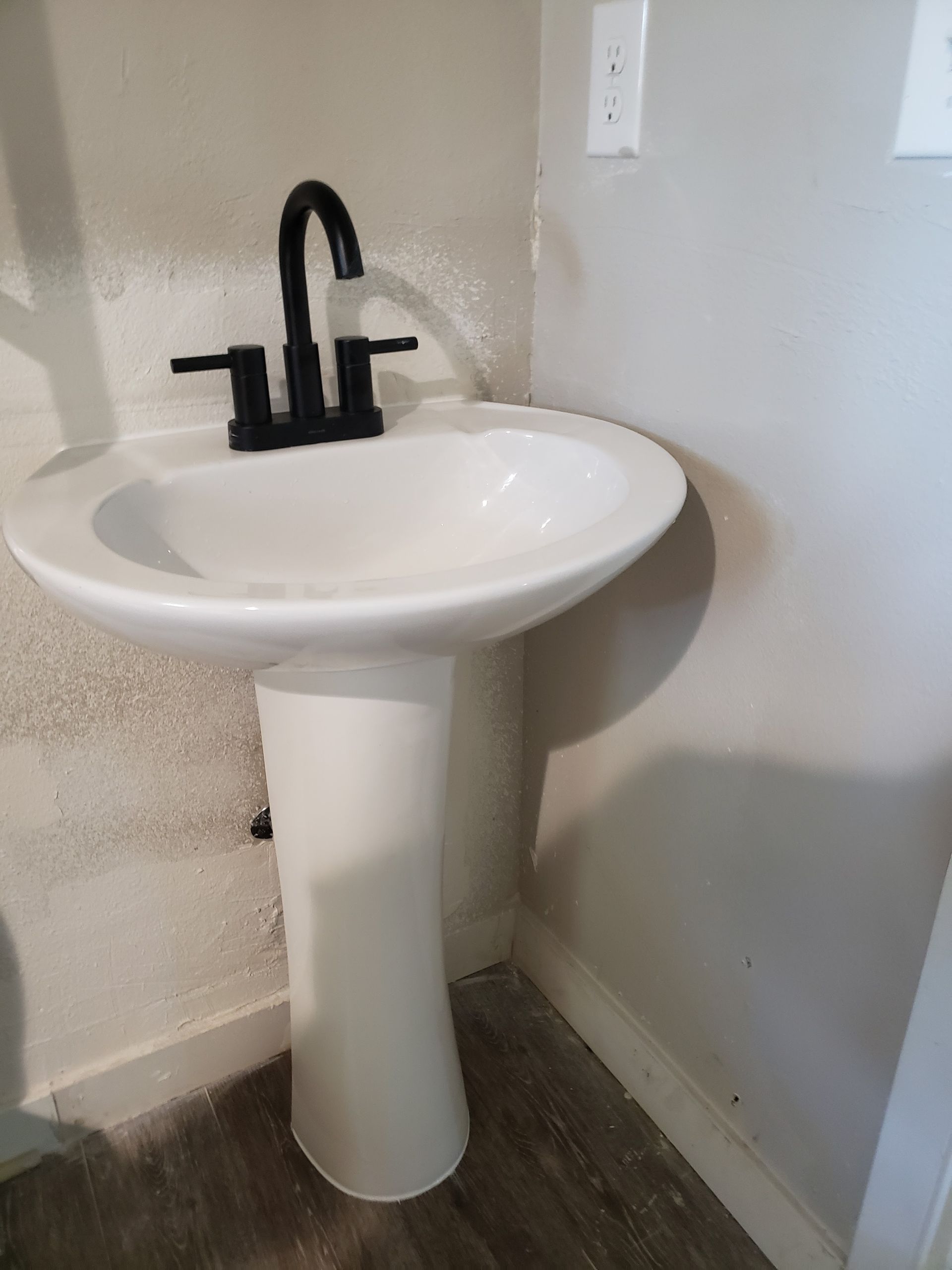 White pedestal sink with black faucet in a small corner bathroom.