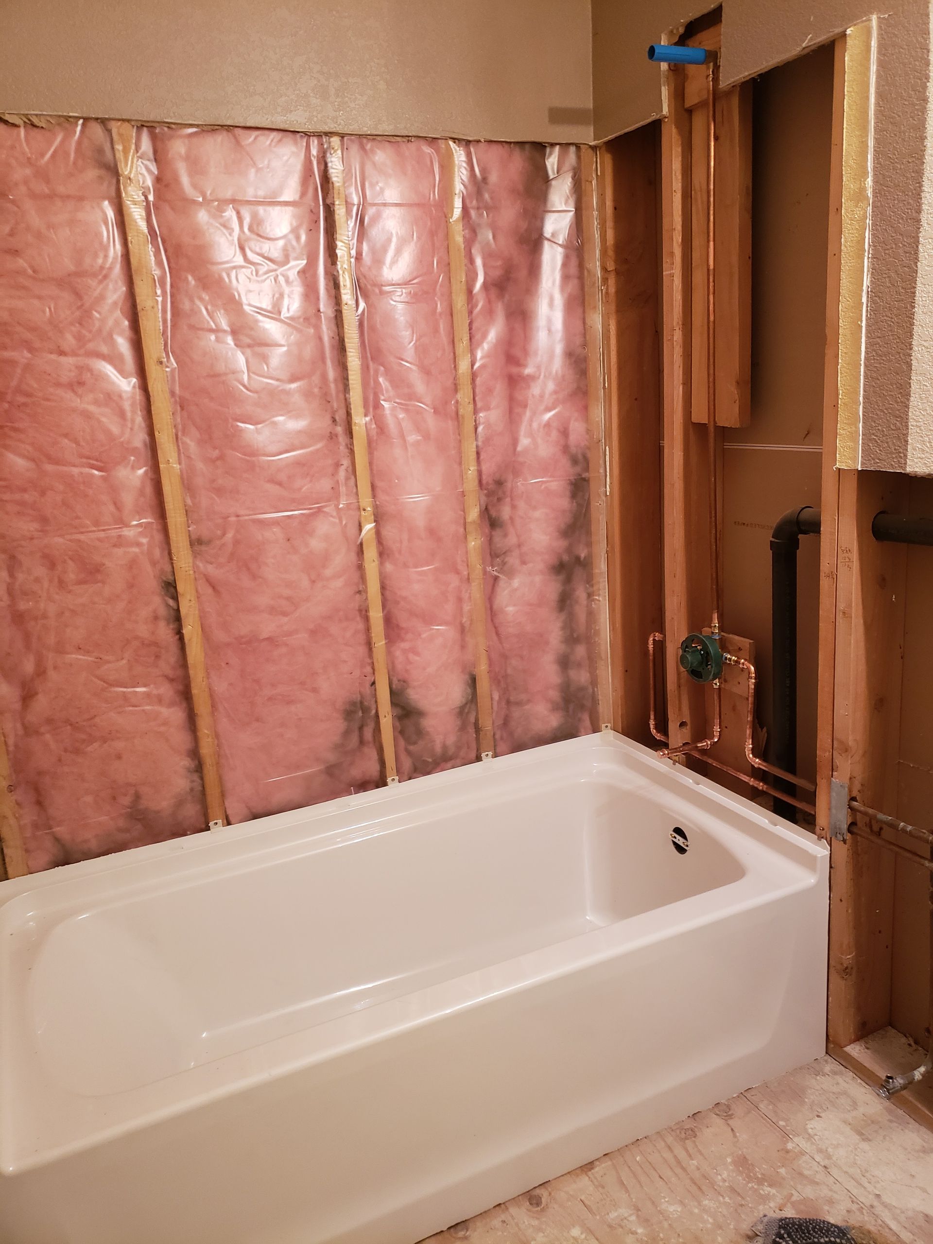 Bathroom under renovation with pink insulation exposed behind a white bathtub and unfinished wood framing