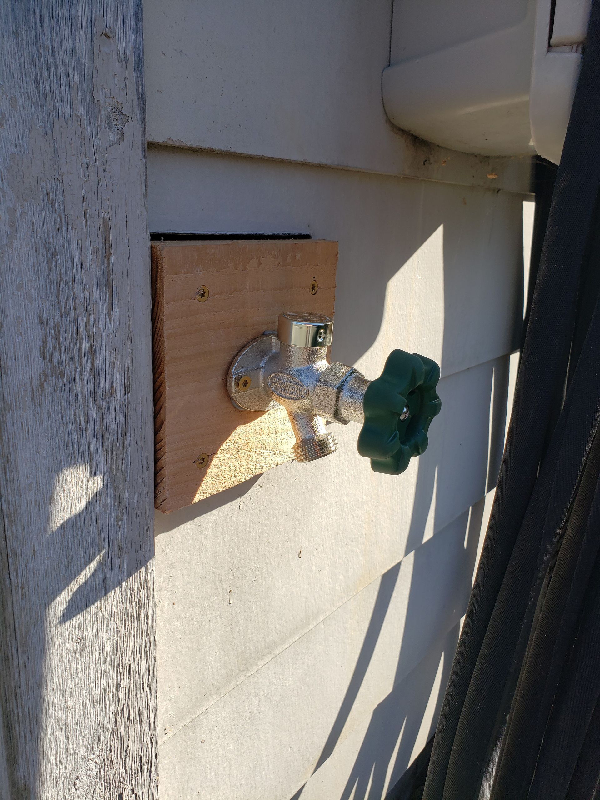 Outdoor water spigot with green handle mounted on a wall beside a wooden board