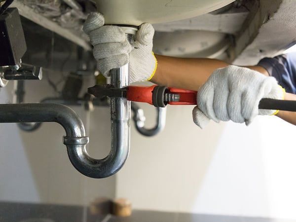 Plumber tightening a pipe connection under a sink with a red wrench, wearing white gloves.