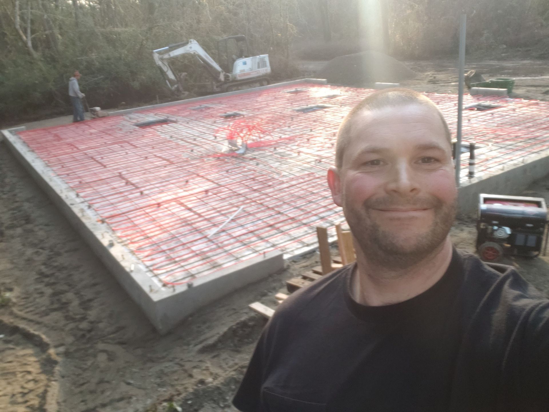 Man smiling in foreground at a concrete foundation under construction, covered with red insulation grid.