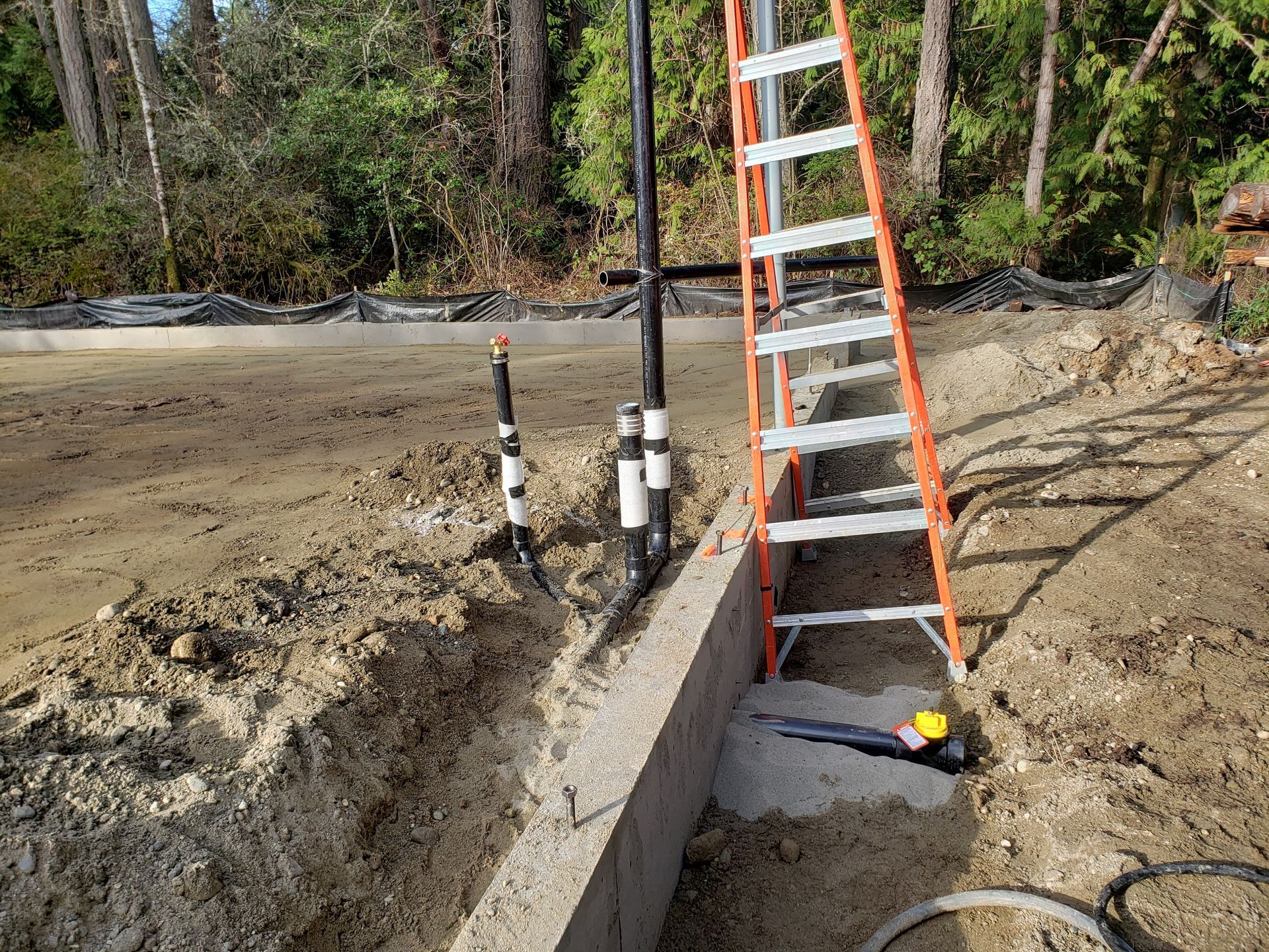 Orange ladder beside a concrete retaining wall at a muddy construction site with pipes and trees in the background