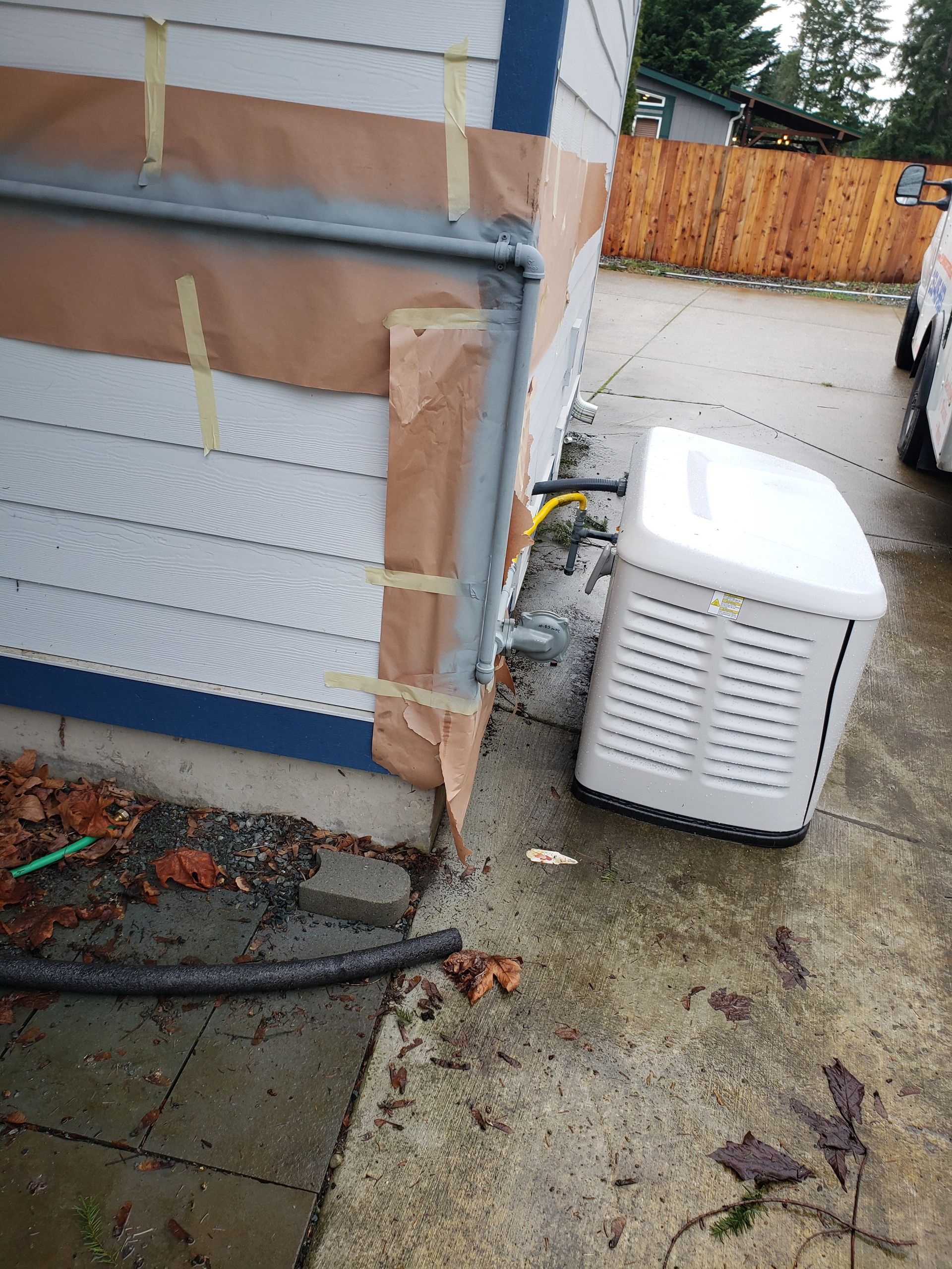 Outdoor AC unit beside a house wall on a wet patio, with a hose and fallen leaves nearby.