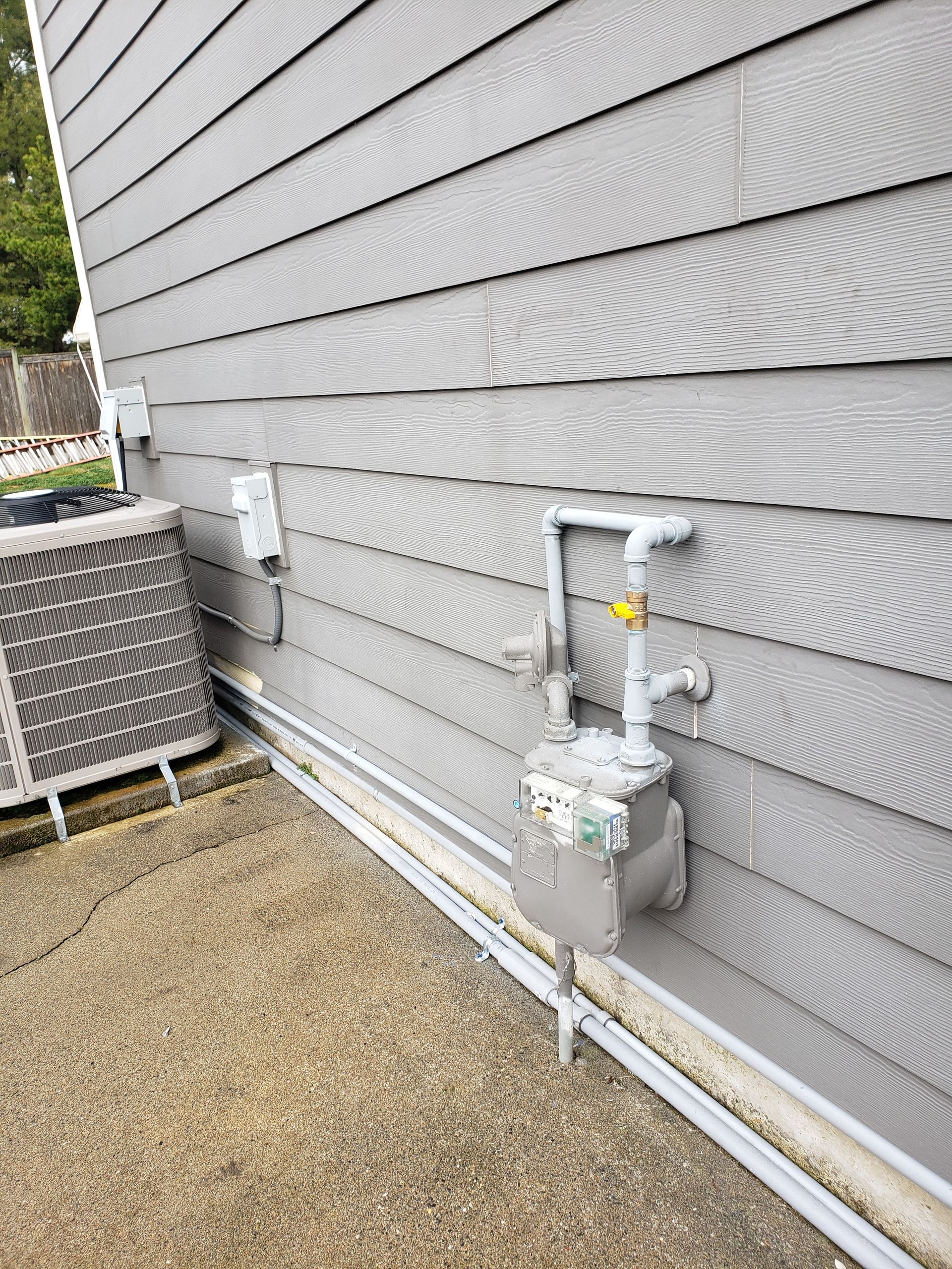 Gray house siding with utility meters and white pipes along a gravel side yard