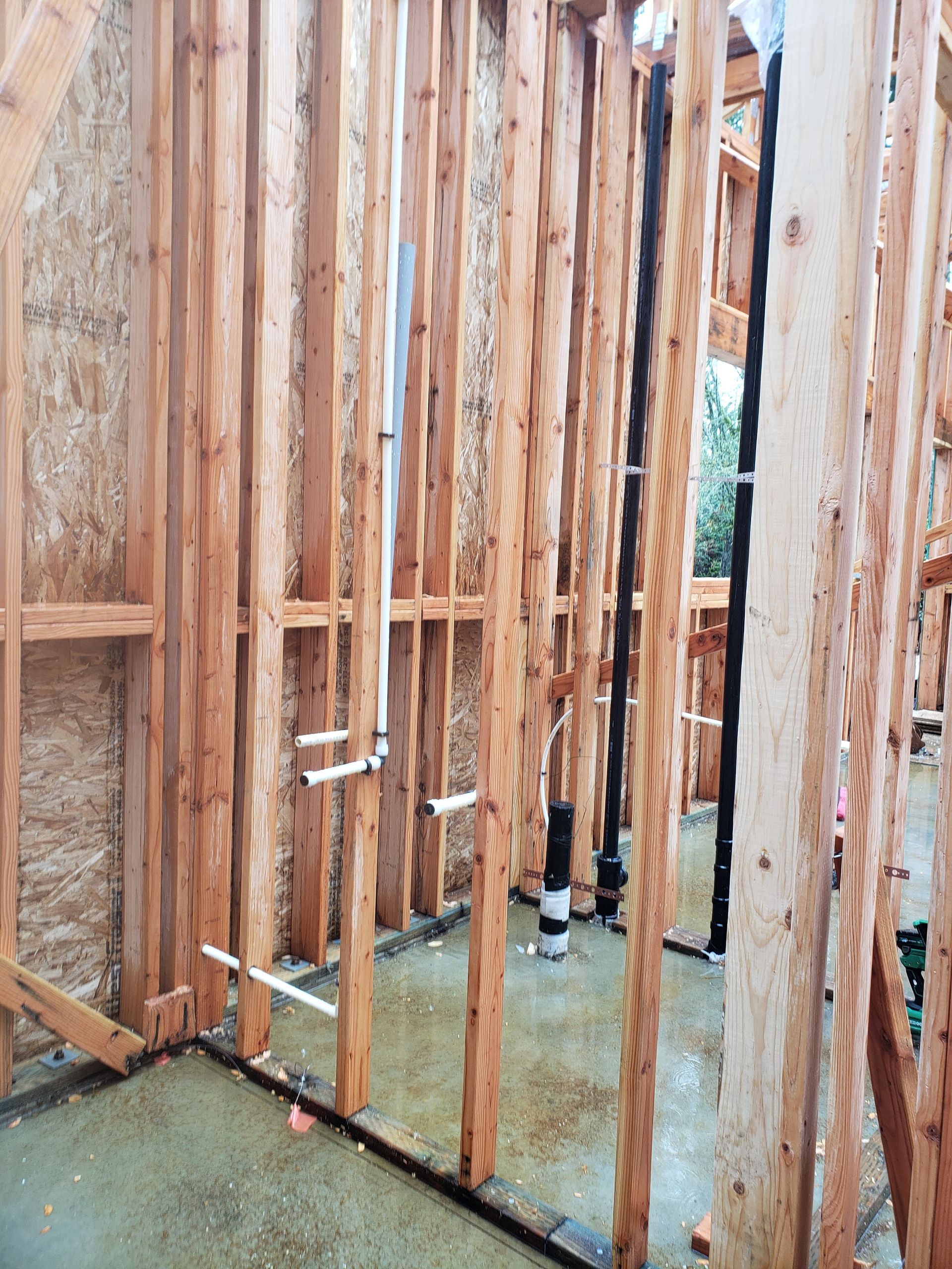 Unfinished wooden wall framing on a concrete floor with exposed studs and plumbing pipes