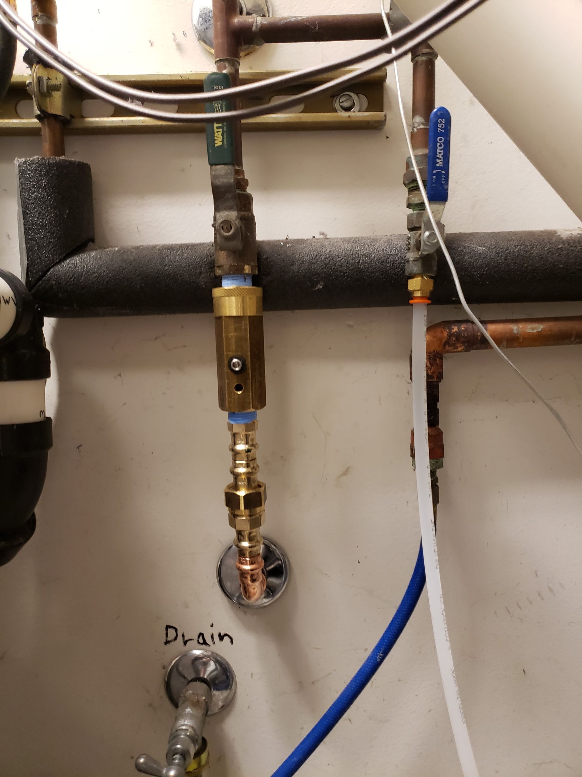 Plumbing pipes and valves against a wall, with a drain labeled “Drain” and a blue hose attached.