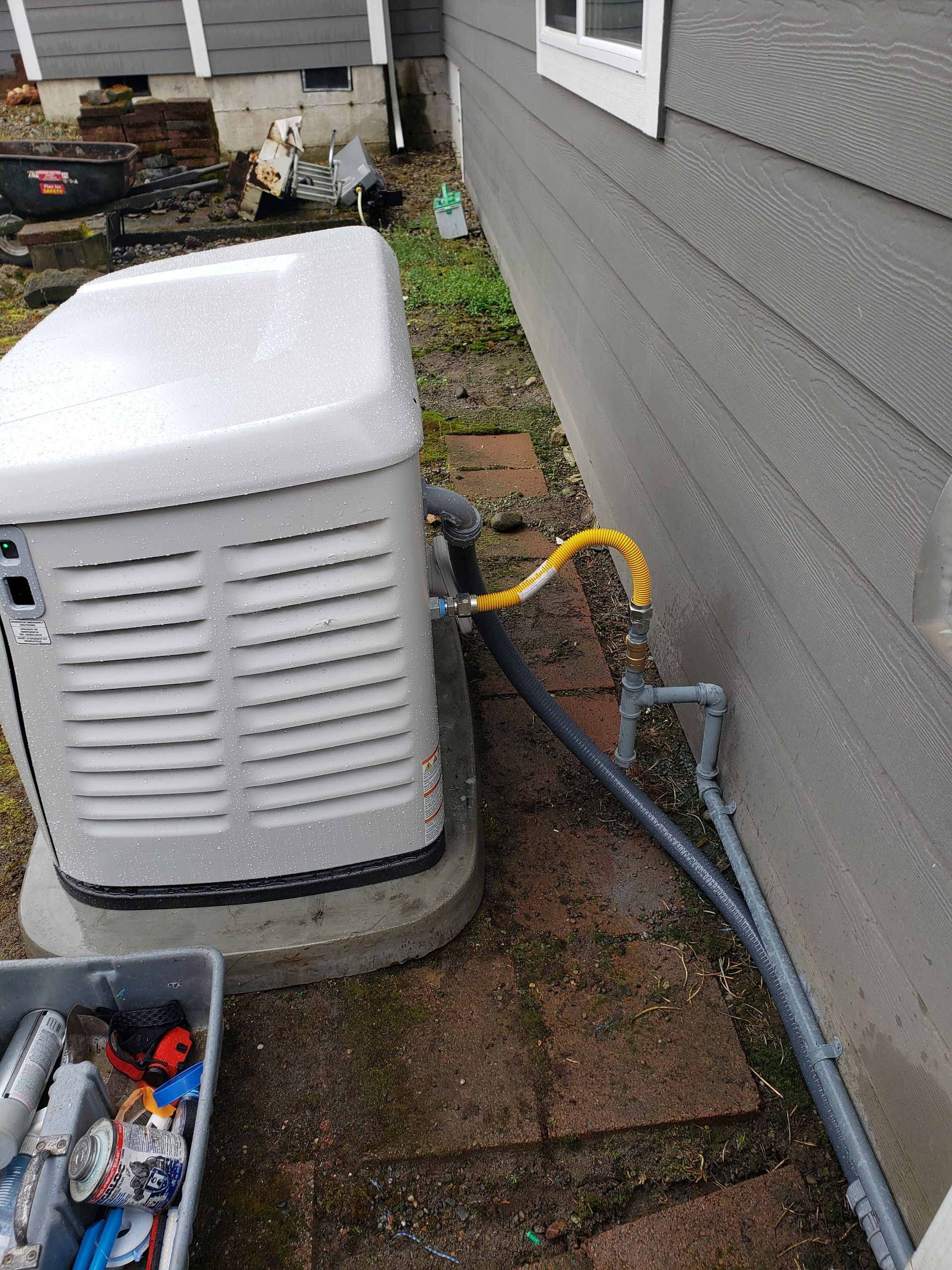 Outdoor HVAC unit beside a gray house, with a yellow hose and black cable running along the wall.