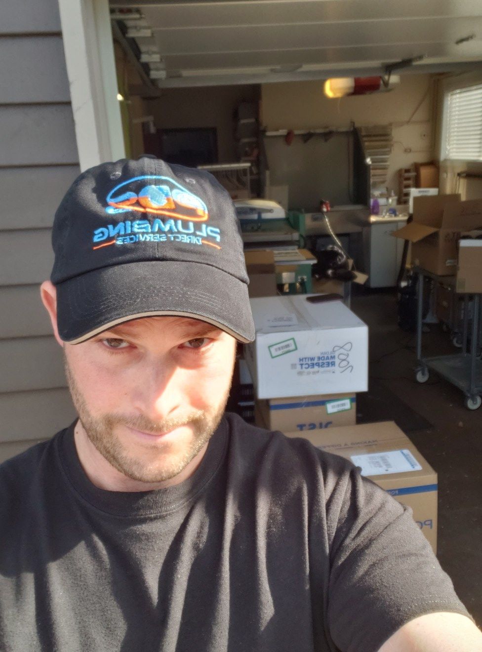 Man taking a selfie in a garage, wearing a black cap and T-shirt, with boxes and shelves behind him