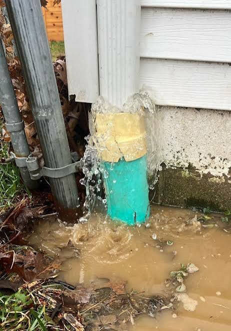 Broken water pipe spraying muddy water beside a house foundation and utility poles.