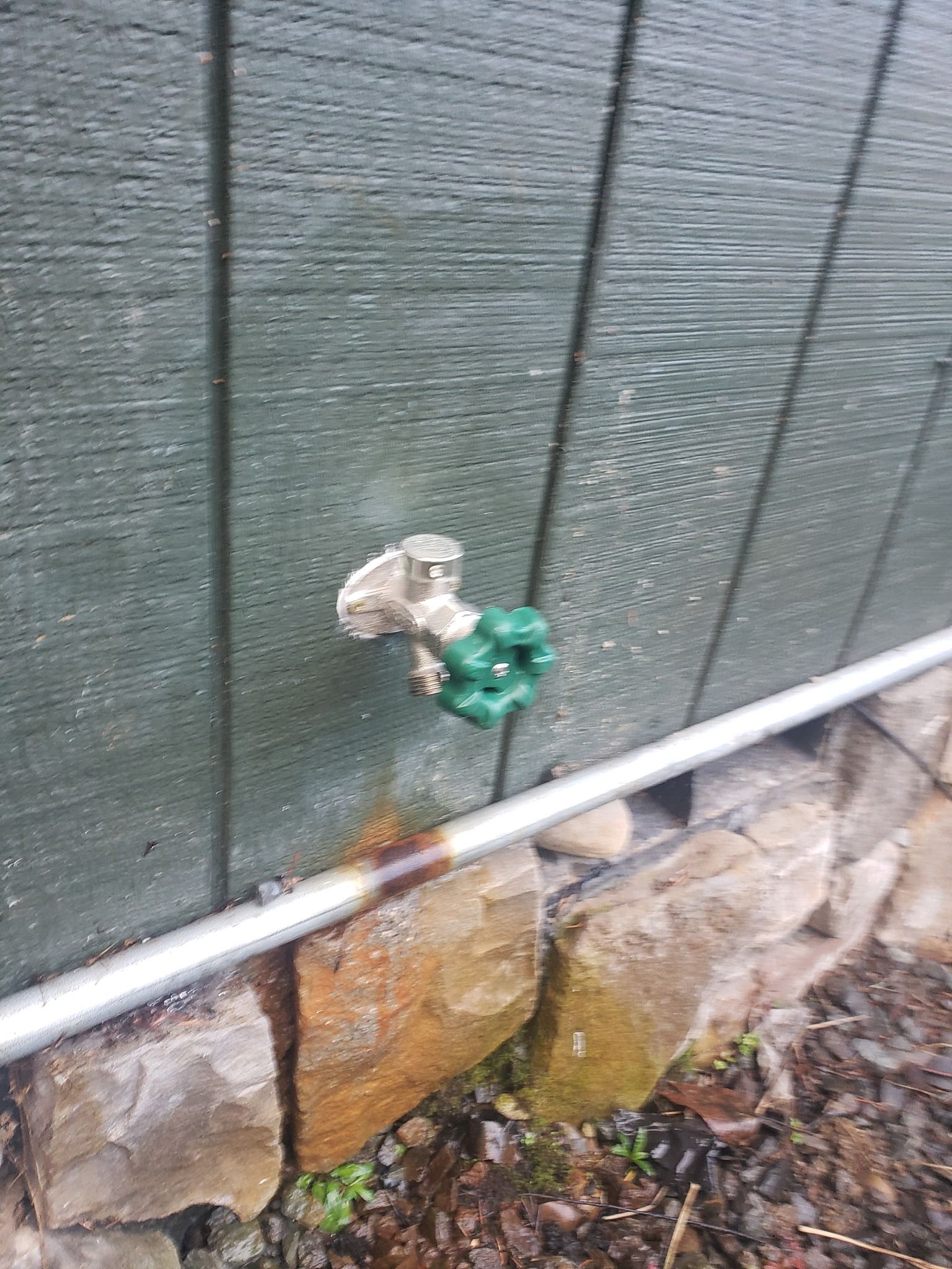 Outdoor faucet with green and silver handles mounted on a green fence above a stone wall.