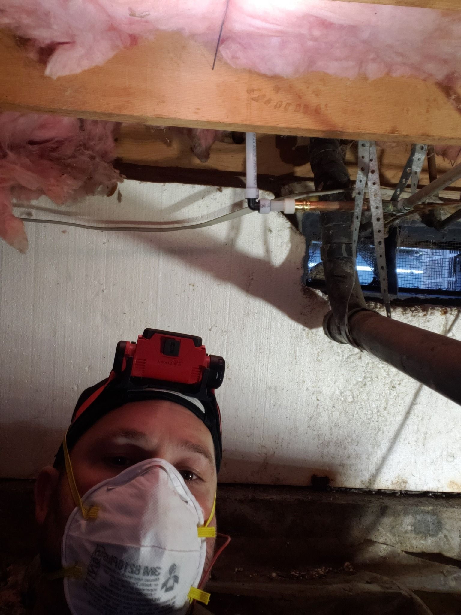 Worker wearing a mask and headlamp inspects exposed ceiling beams and wiring in a crawlspace