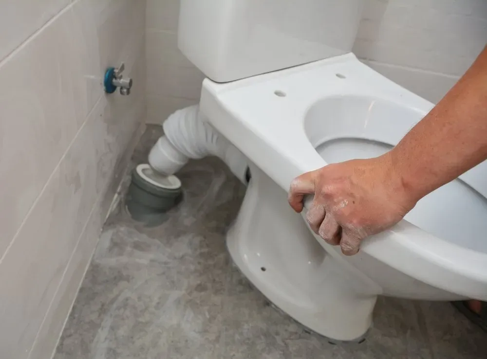 Hand unclogging a toilet with a plunger and gray hose on the floor, water pooled around it