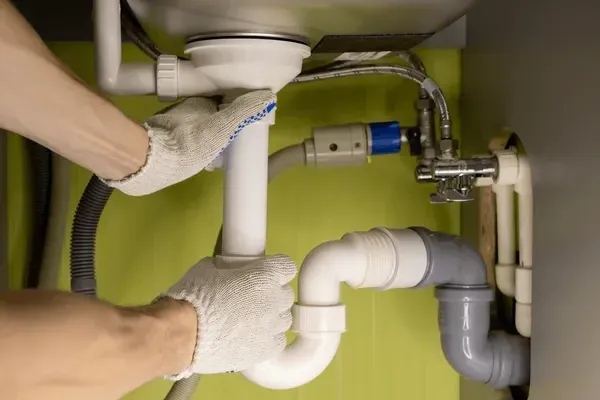 Hands connecting white PVC sink pipes under a green cabinet, with an adjustable wrench visible on the right.