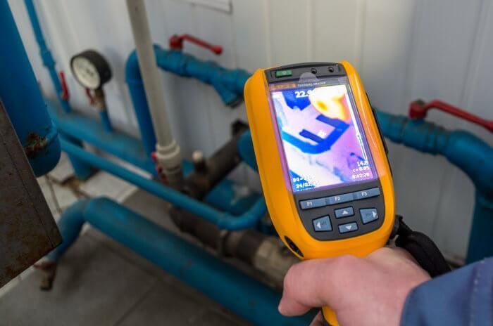 Handheld thermal camera scanning blue industrial pipes and valves in a utility room