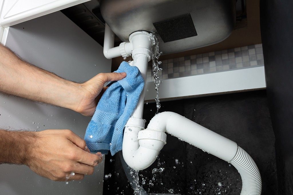 Person cleaning a leaking white sink pipe with a blue cloth over a black bucket