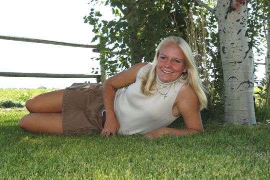 Blond person lying in grass, smiling, wearing tan top and brown shorts near a tree and fence.