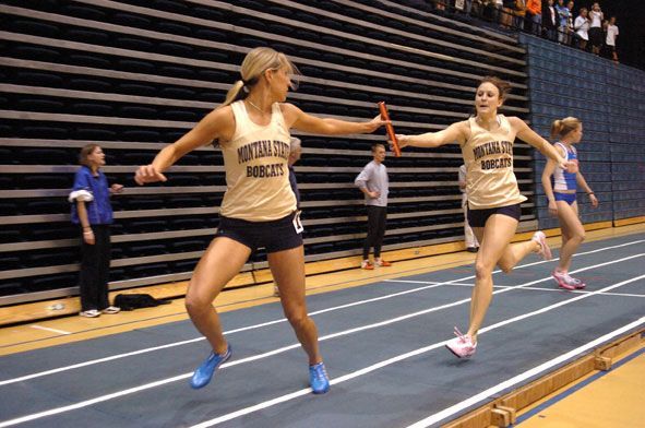 Two runners in matching gold tops and black shorts exchange a baton in an indoor track race.